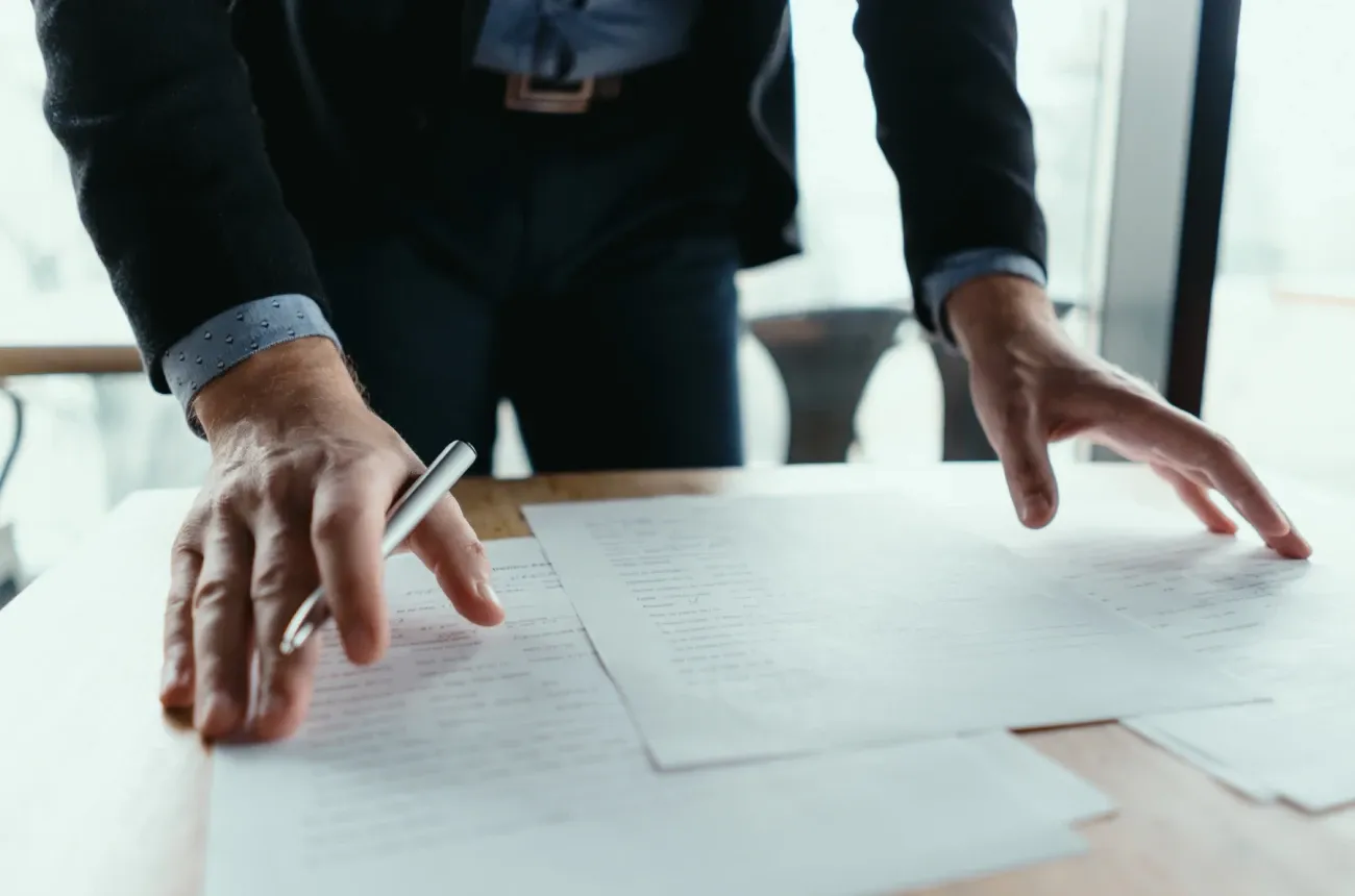 A man in a dark blazer leans over a wooden desk with a pen in hand, reviewing printed documents