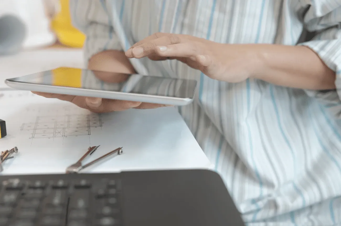 A person in a striped shirt using a tablet over building blueprints, with drafting tools and a laptop on the desk