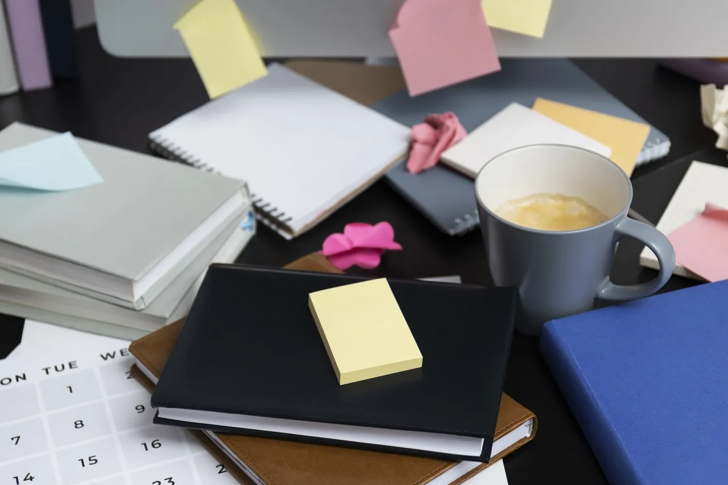 A cluttered desk covered in notebooks, sticky notes, a coffee mug and a paper calendar