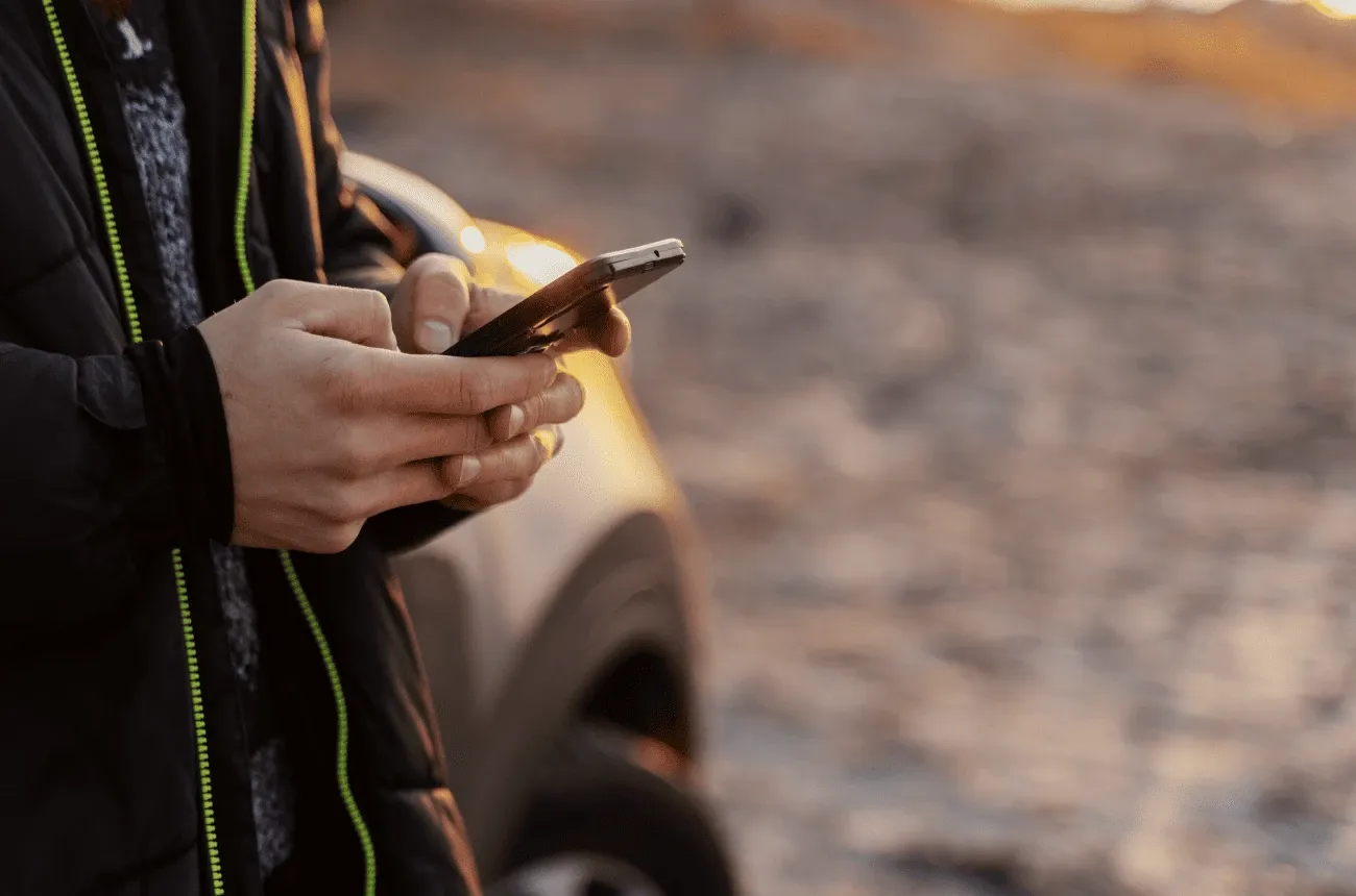 A person in a black jacket typing on a smartphone next to a parked car at sunset