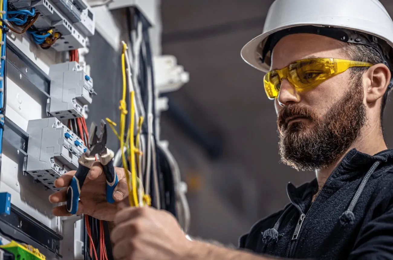An electrician in a white hard hat and yellow safety glasses stripping wires at an open electrical panel