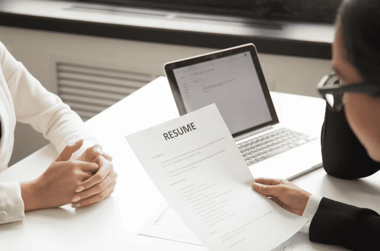 A person holds a printed resume across a desk from a job candidate, with a laptop open between them