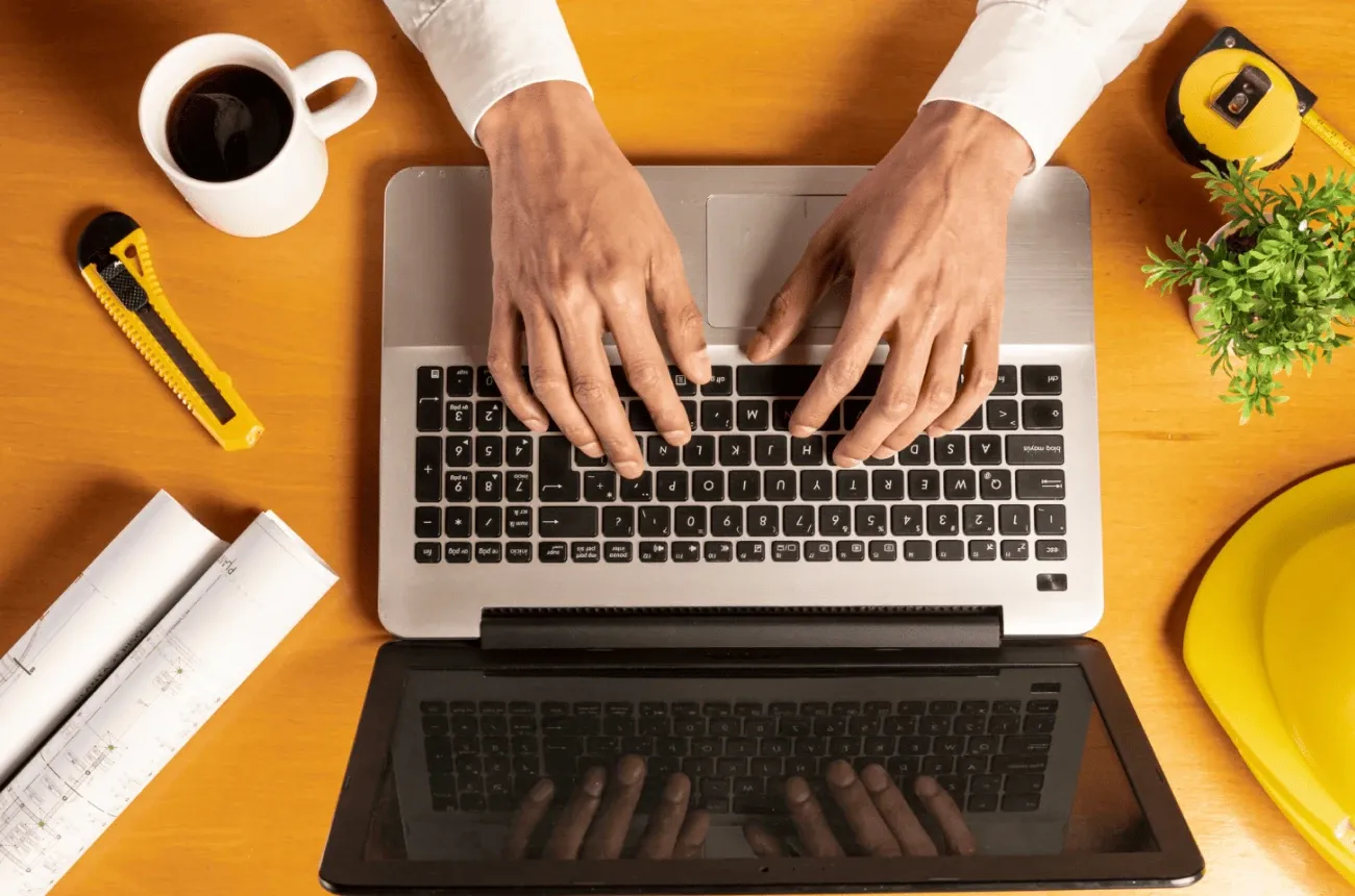 Overhead view of hands typing on a laptop on a desk with coffee, blueprints, a tape measure and a hard hat