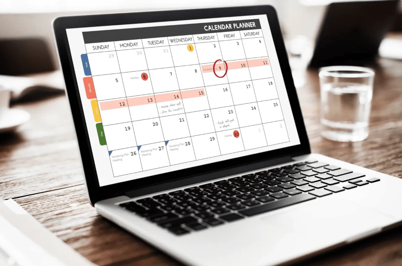 A laptop on a wooden table showing a monthly calendar planner, next to a glass of water