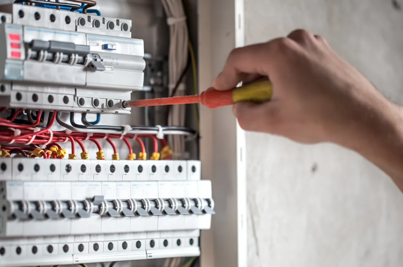 A hand using a screwdriver on circuit breakers and red wiring inside an electrical panel