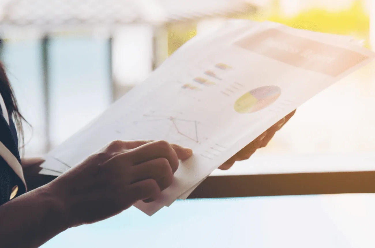 Hands holding printed business report pages with a pie chart and a line graph next to a bright window