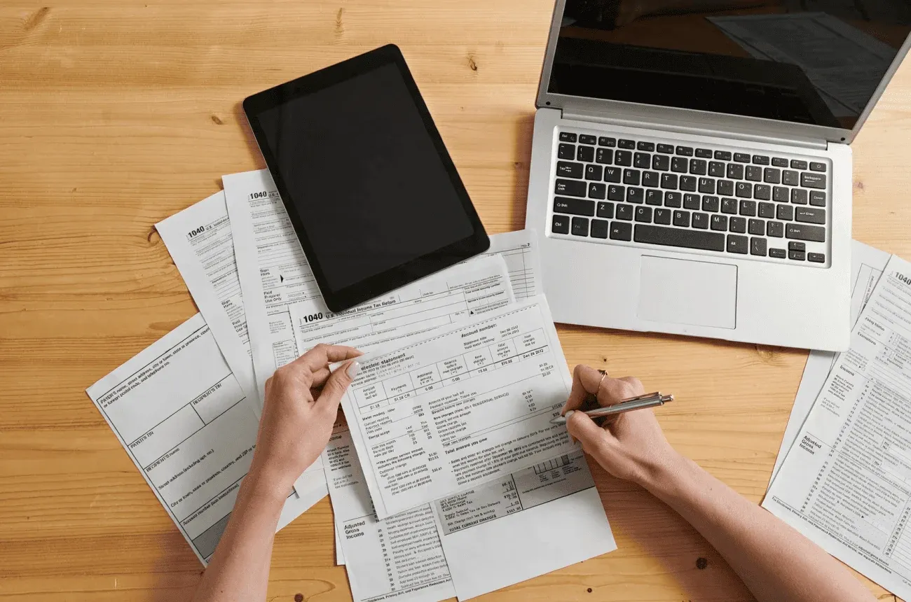 Hands checking an electric bill with a pen among tax forms, next to a tablet and a laptop on a wooden desk
