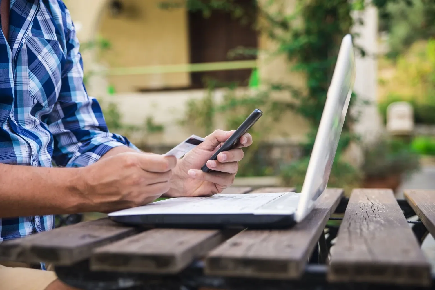 A person in a plaid shirt holding a payment card and a smartphone at a laptop on an outdoor wooden table