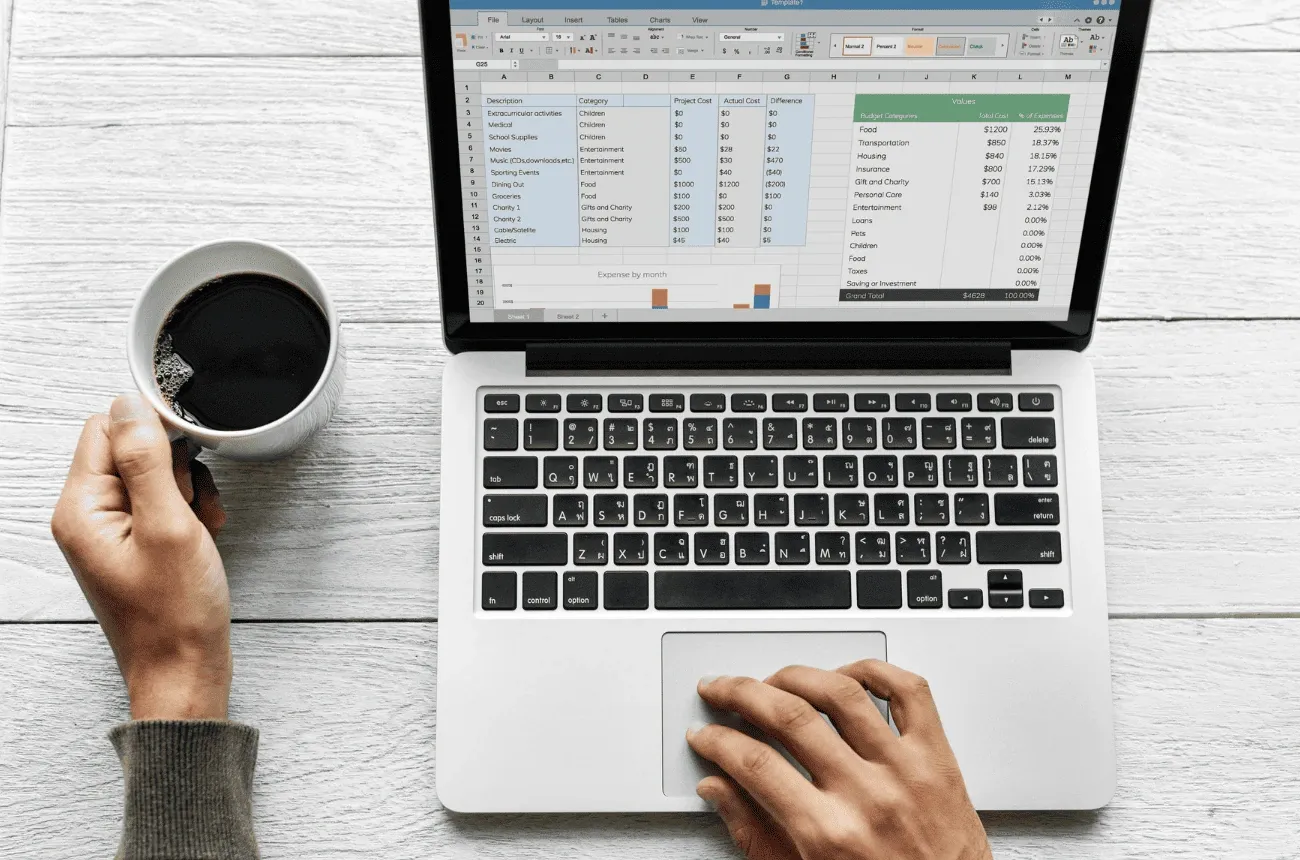 A person holding a coffee mug while using a laptop with a spreadsheet open on a white wooden table