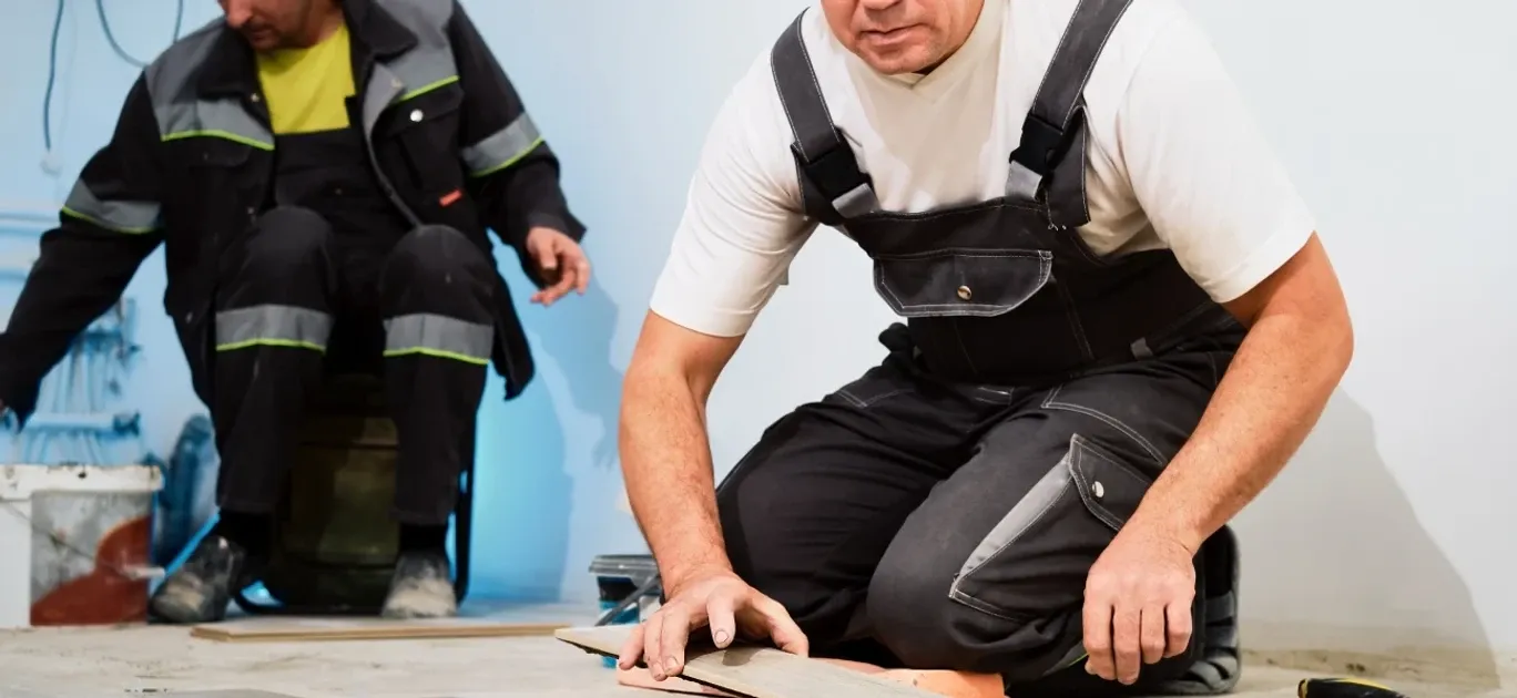A worker in black overalls kneels to lay a floor plank while a coworker in reflective workwear sits behind him