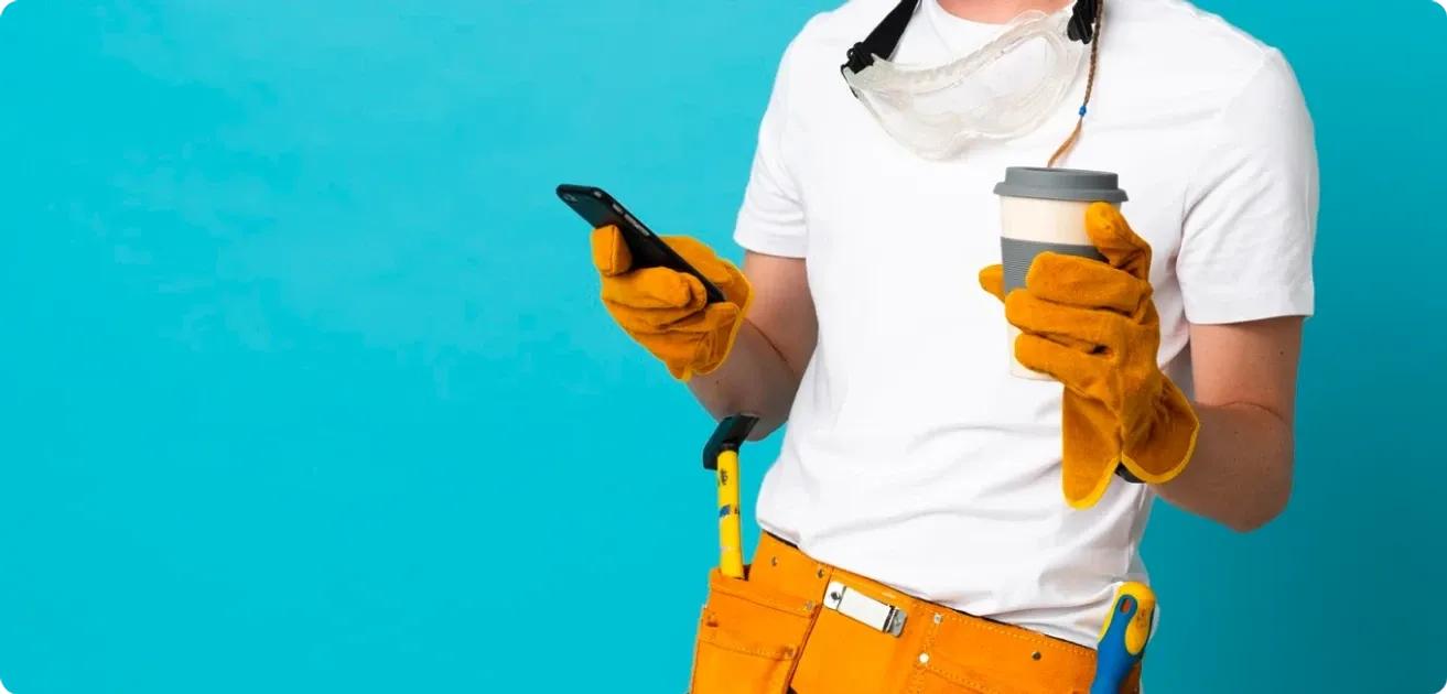 A worker in work gloves, safety goggles and a tool belt holding a phone and a coffee cup against a blue background