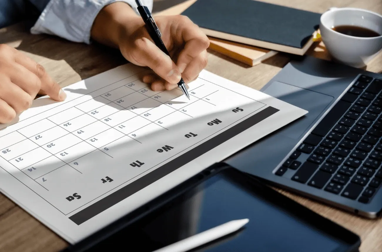 A person writes on a printed monthly calendar with a pen next to a laptop, a tablet and a cup of coffee
