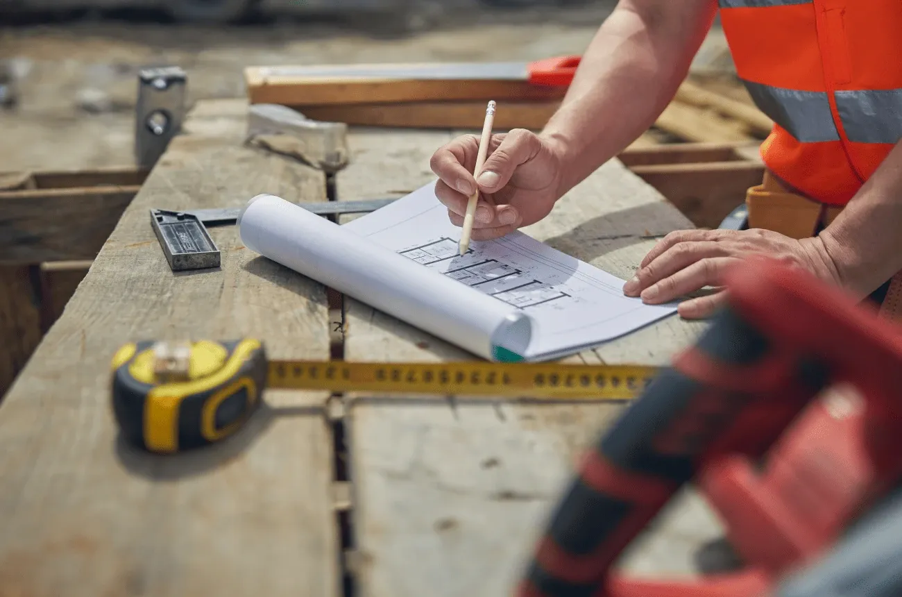 A worker in an orange safety vest marking a floor plan with a pencil on wooden boards next to a tape measure