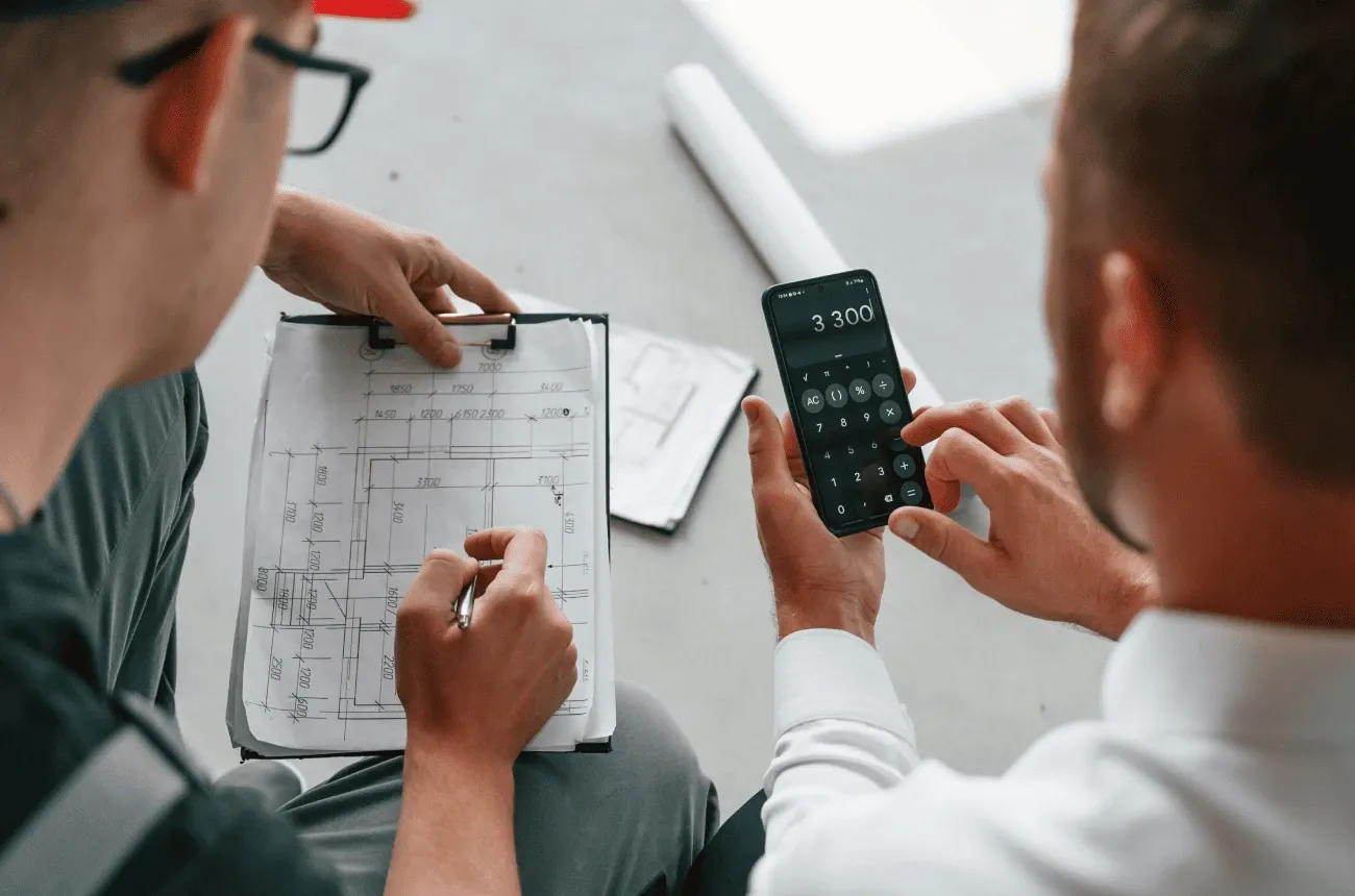 Two men review a floor plan on a clipboard while one totals figures on a phone calculator
