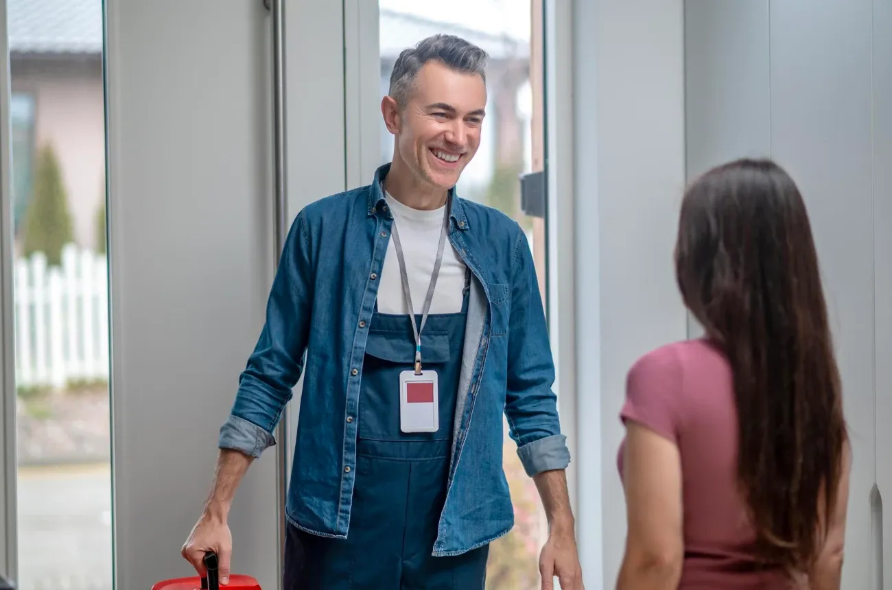 A smiling repairman in a denim shirt and overalls with a red toolbox greets a woman at the front door