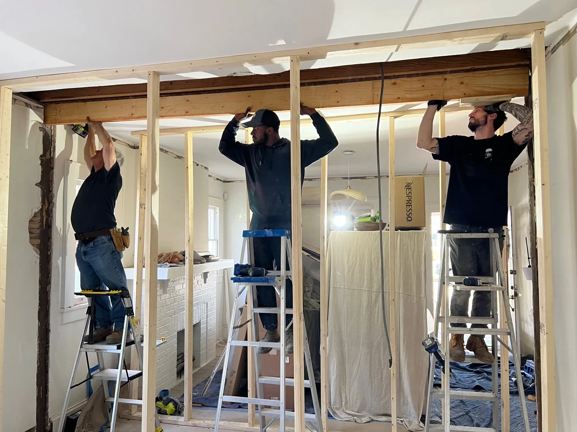 Three workers on ladders fitting a wooden header beam into a newly framed wall during a home remodel