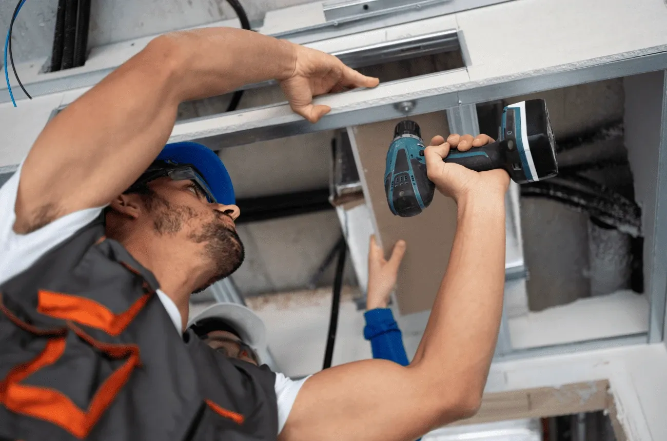 A worker in a blue hard hat uses a cordless drill on a ceiling vent while a coworker helps