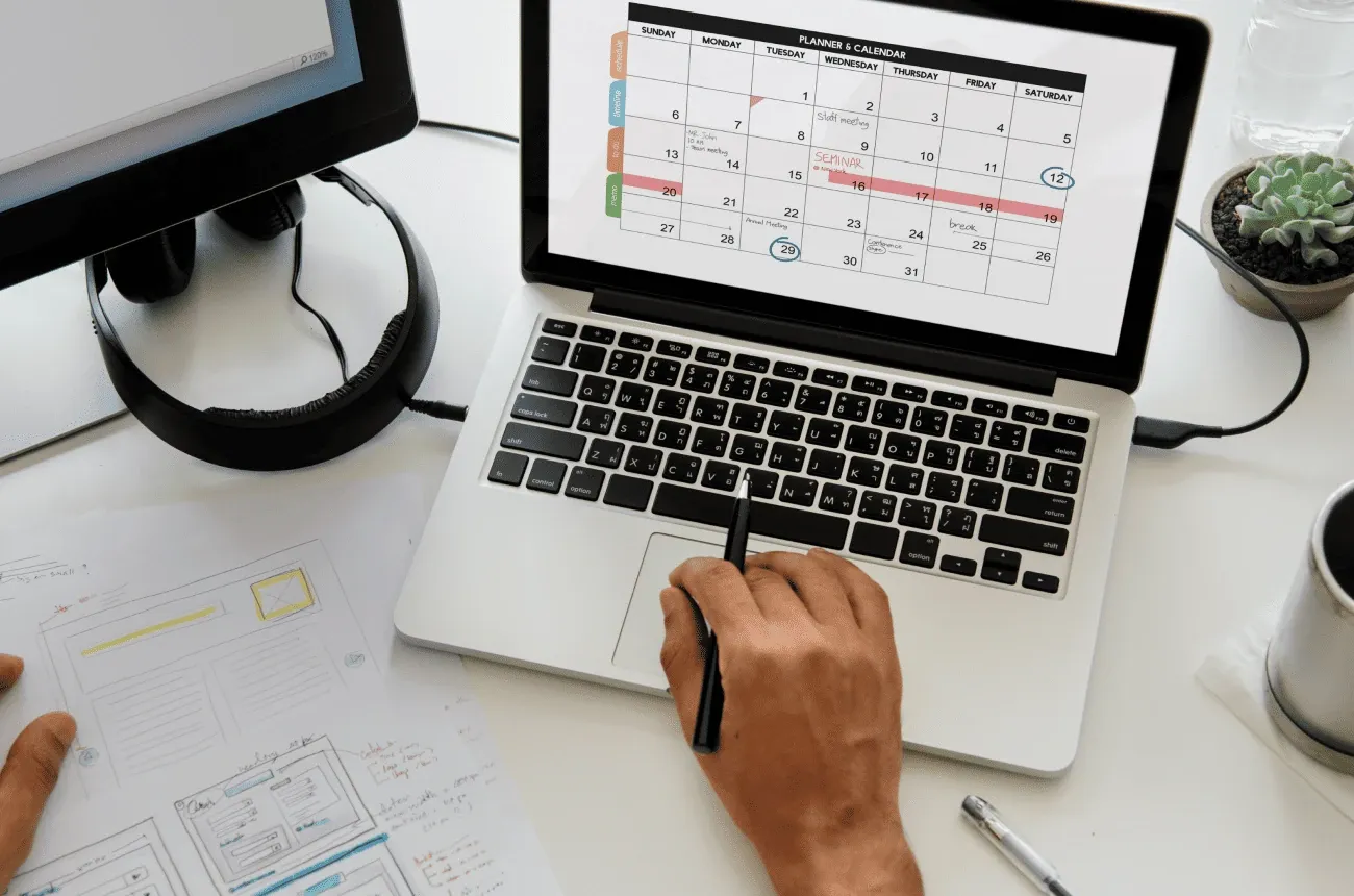 A person holding a pen over a laptop showing a monthly planner calendar, with wireframe sketches on the desk
