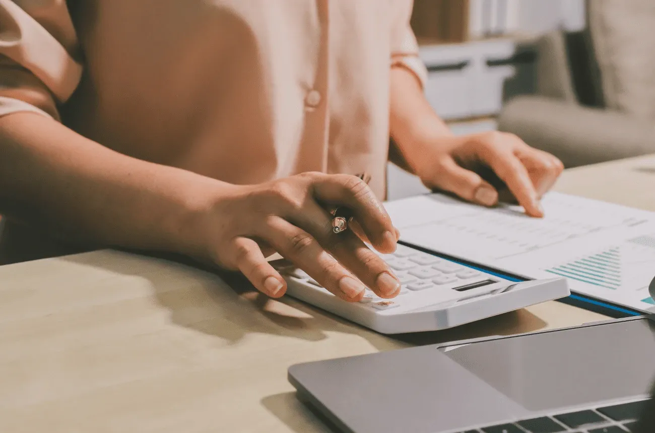 Hands using a white calculator with a pen next to printed charts and a laptop on a wooden desk