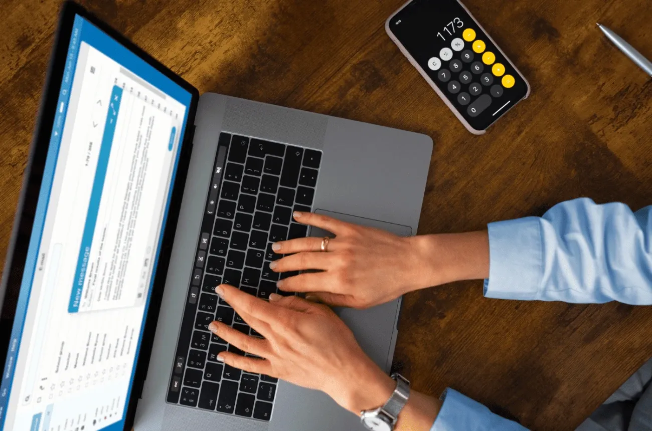 Hands typing on a laptop on a wooden desk next to a phone showing a calculator app