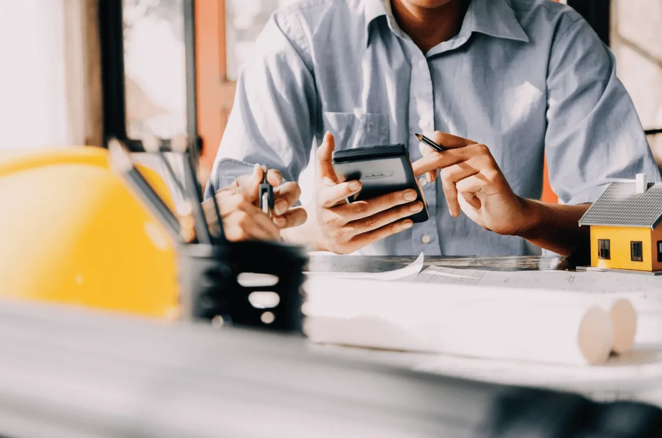 A person in a blue shirt holding a calculator and pencil at a desk with blueprints, a hard hat, and a small house model