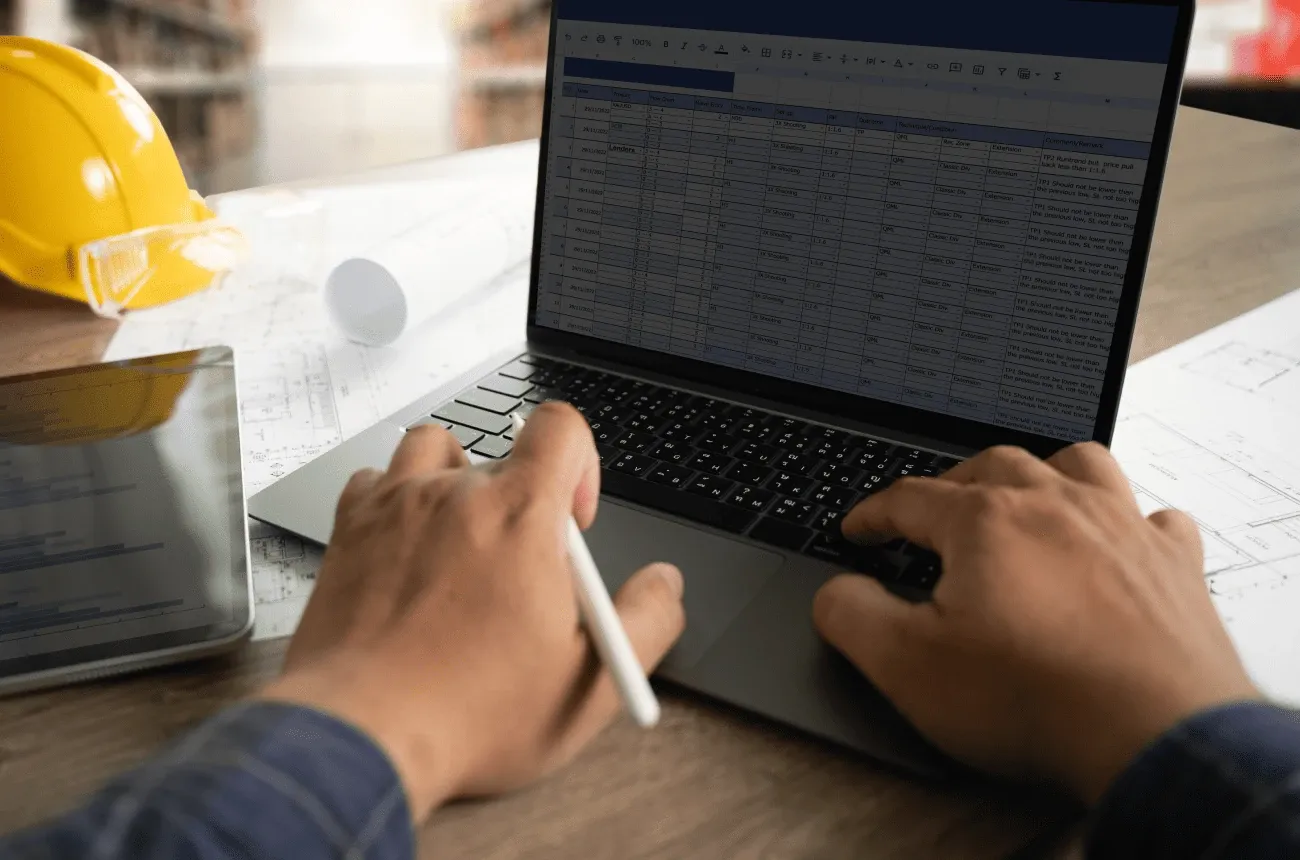 Hands typing on a laptop showing a spreadsheet, next to a yellow hard hat, a tablet and blueprints on a desk