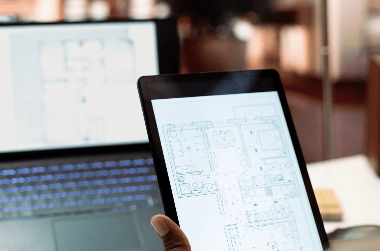 A hand holding a tablet showing an apartment floor plan in front of a laptop displaying another plan
