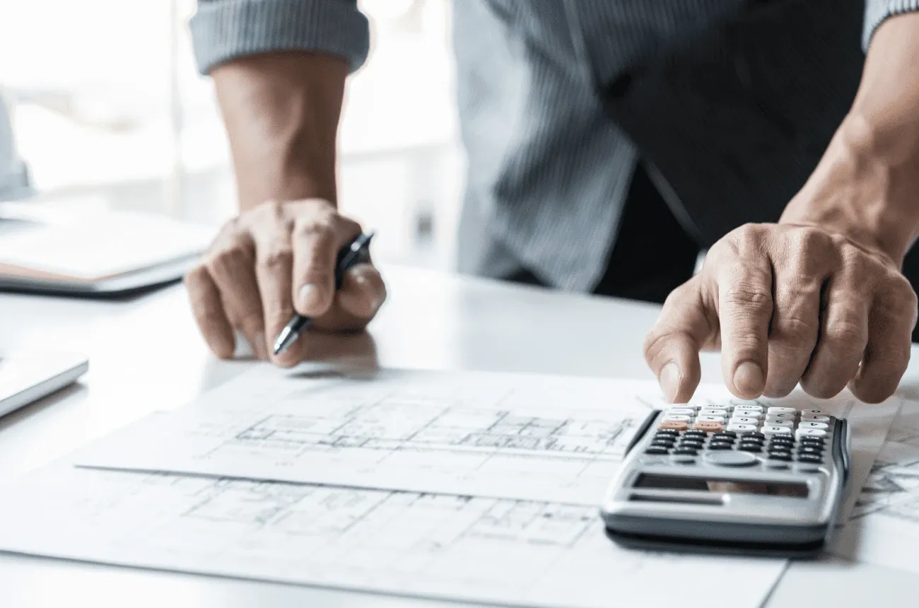 A person holding a pen taps a calculator over building plans on a white desk