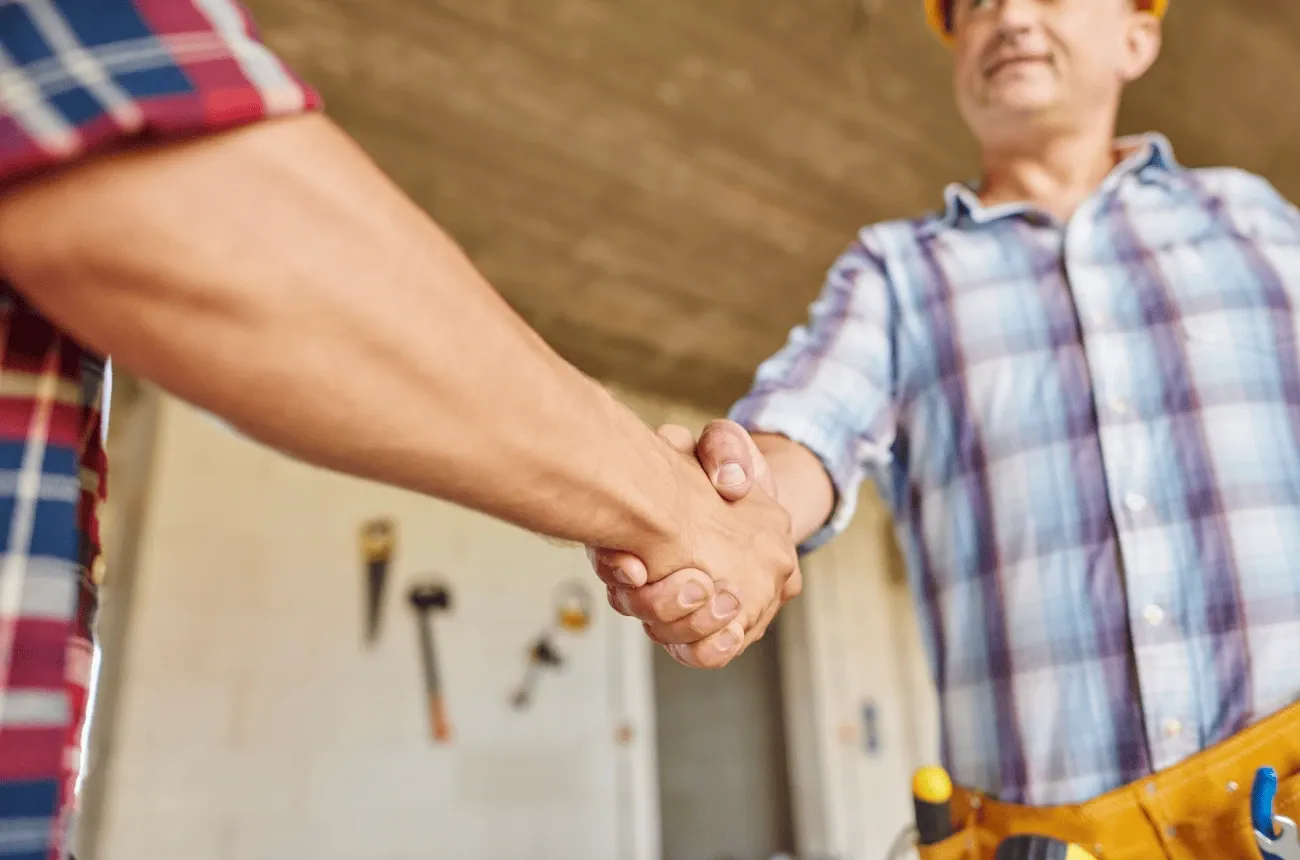 A contractor in a hard hat and tool belt shakes hands with a man in a plaid shirt at a work site