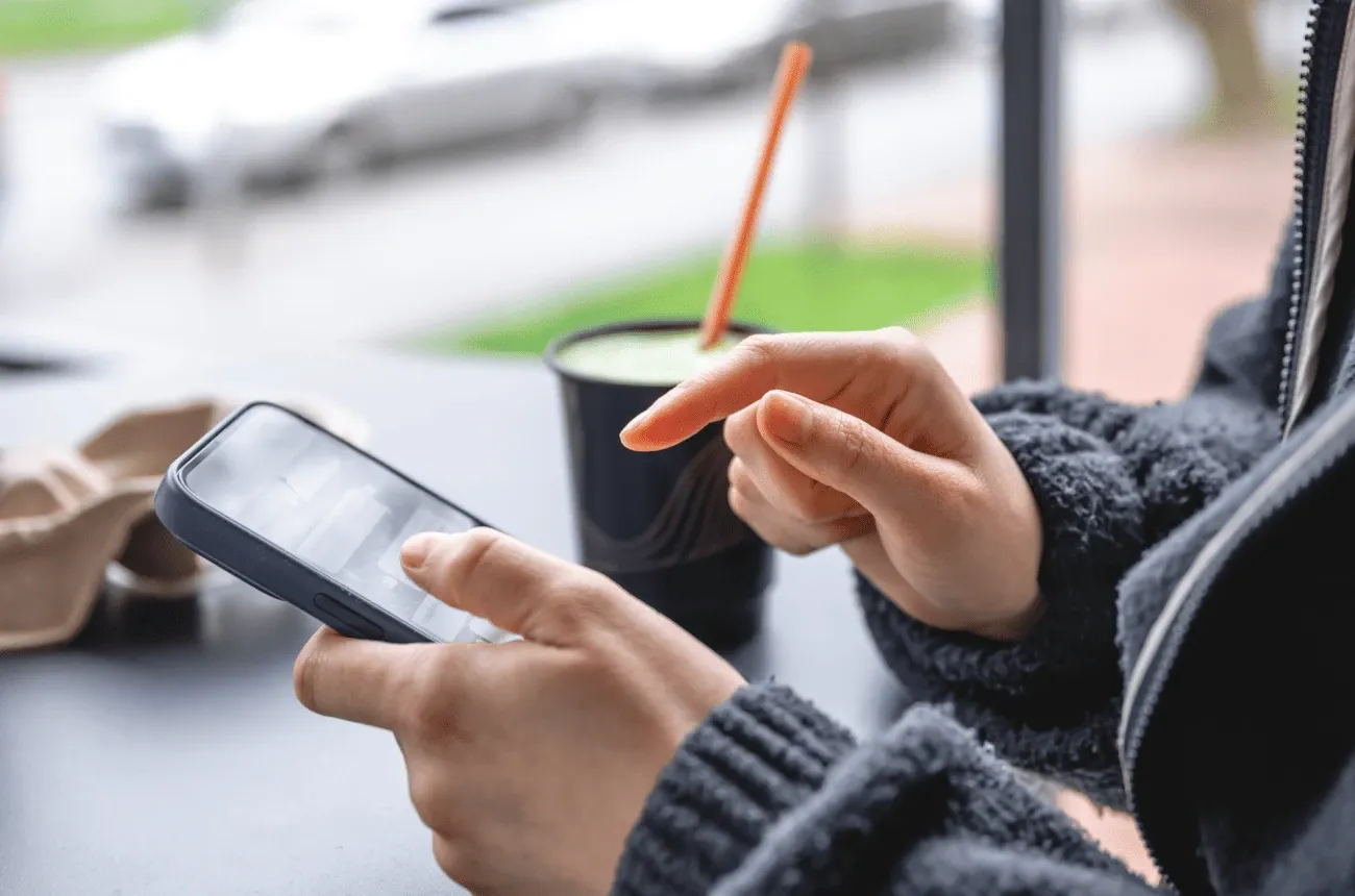 A person in a gray sweater using a smartphone at a table next to a cup with an orange straw