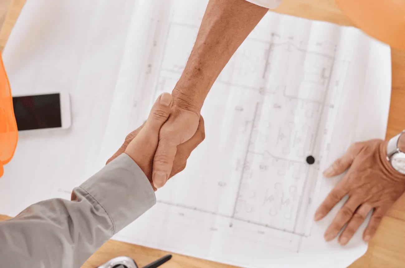 Two people shake hands over a building floor plan on a table next to a phone