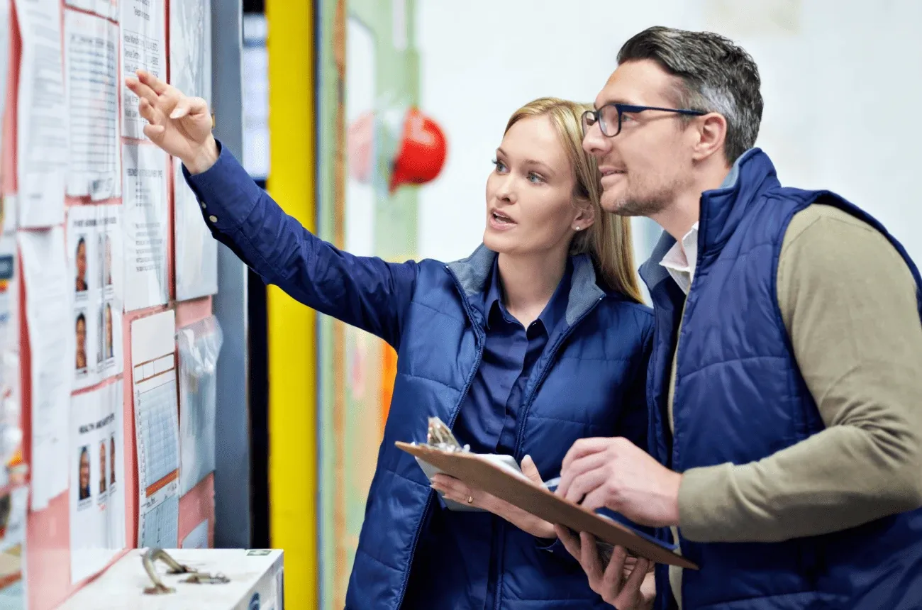 Two coworkers in navy vests look at papers pinned to a wall board, one pointing while the other holds a clipboard