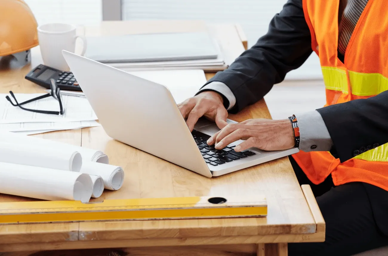 A person in a suit and orange safety vest types on a laptop at a desk with rolled blueprints, a level and a hard hat
