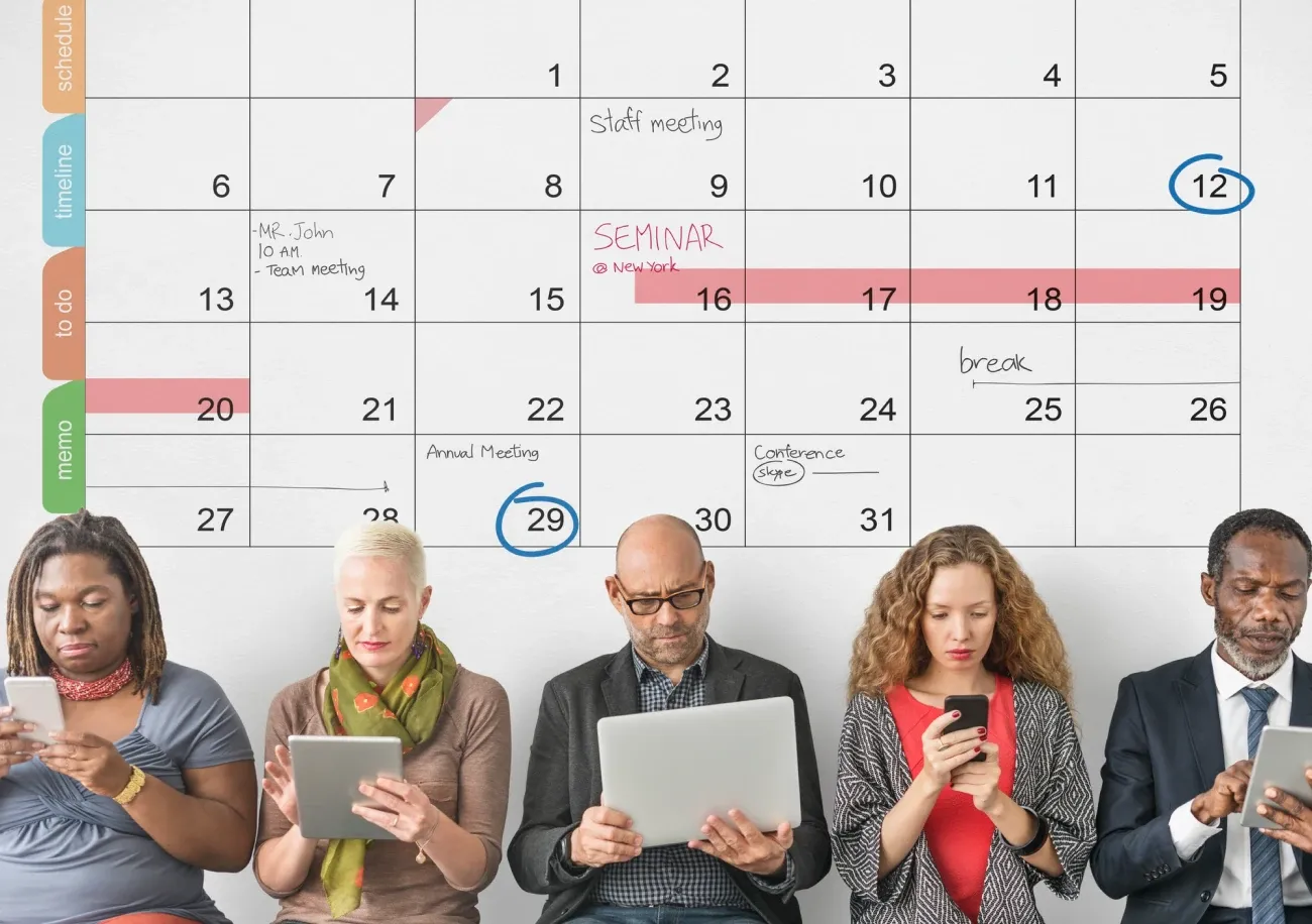 Five people using phones, tablets and a laptop in front of a large wall calendar with notes and circled dates