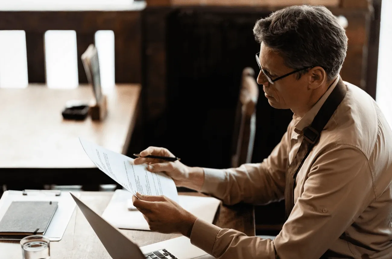 A man with glasses and a beige shirt reviewing printed documents with a pen at a wooden table with a laptop