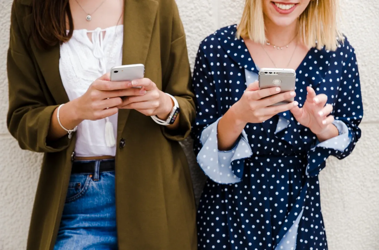 Two smiling women in casual clothes leaning against a wall and looking at their smartphones