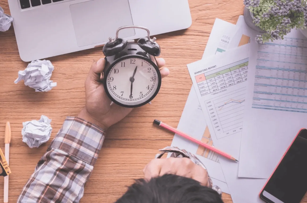 A person holding an alarm clock over a desk with a laptop, crumpled paper, a pencil, and printed reports