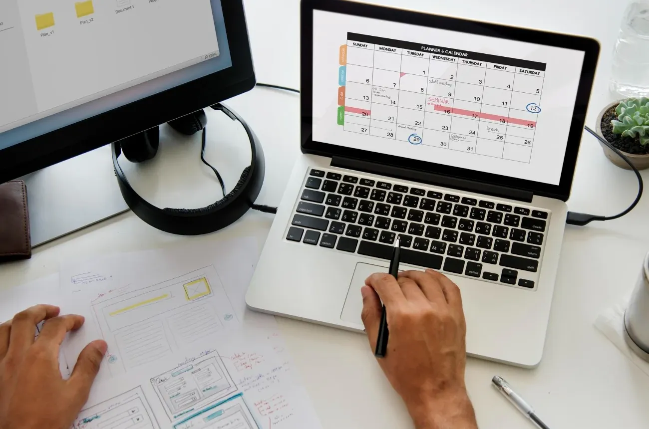 A person pointing a pen at a monthly planner calendar on a laptop, with wireframe sketches on the desk