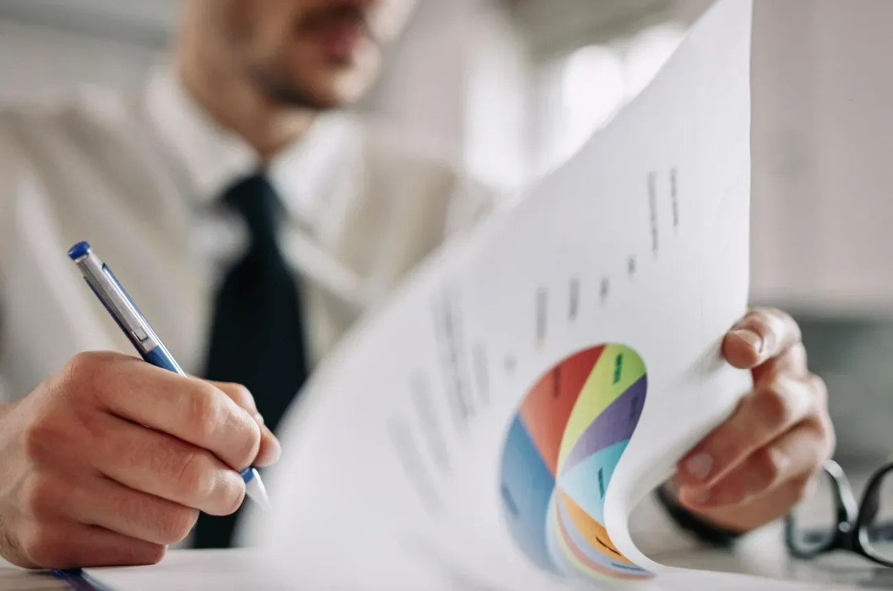 A man in a shirt and tie holding a report with a colorful pie chart while writing with a blue pen
