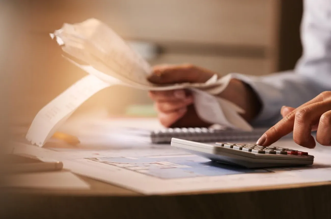 A person holds a stack of paper receipts and types on a calculator at a desk