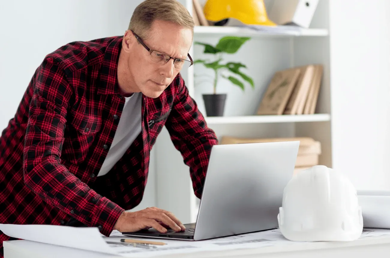 A man in a red plaid shirt and glasses works on a laptop at a desk with a white hard hat and blueprints