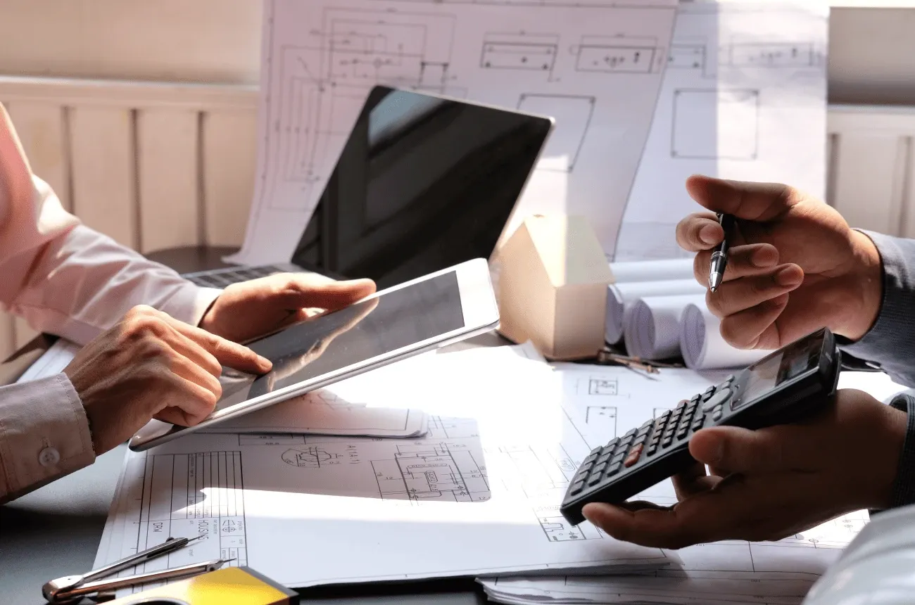 Two people review blueprints at a desk, one using a tablet and the other holding a calculator and a pen