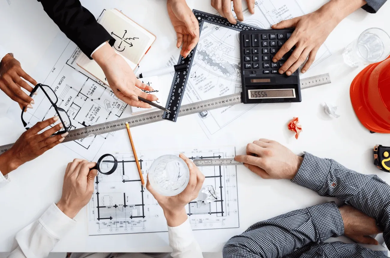 Overhead view of several people’s hands over floor plans with a calculator, rulers, a magnifying glass and a hard hat