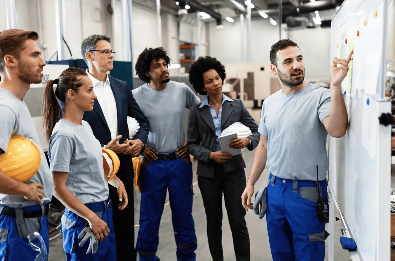 A worker points at a whiteboard while a team in work uniforms and hard hats listens inside a factory