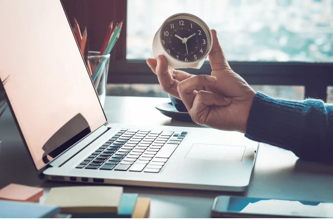 A hand holds a small alarm clock beside an open laptop on a desk with sticky notes and pencils