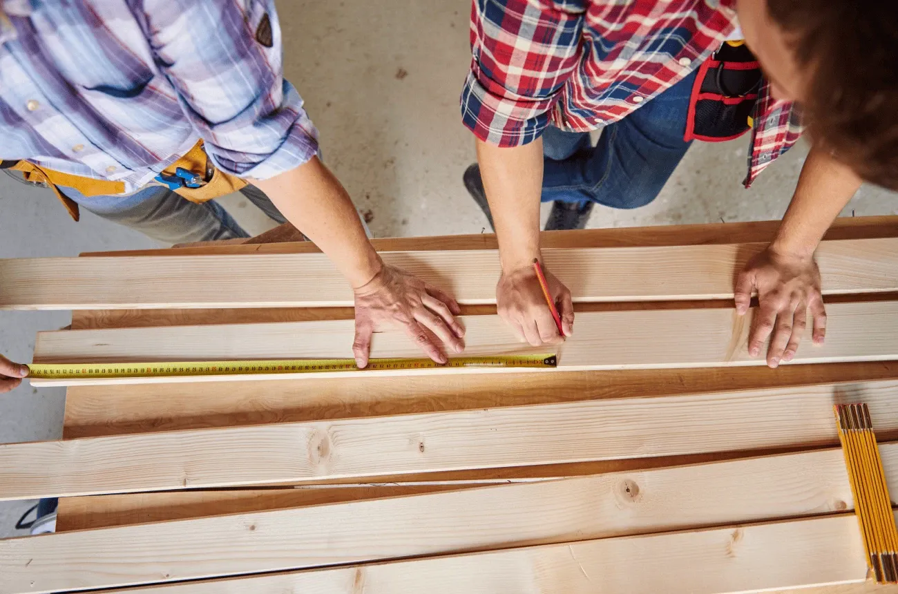 Two workers in plaid shirts measuring and marking wooden boards with a tape measure and a pencil