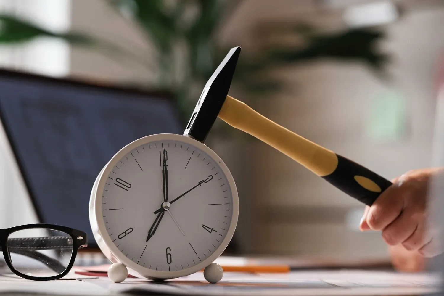 A hand holds a hammer against a white alarm clock on a desk next to a pair of glasses and a laptop