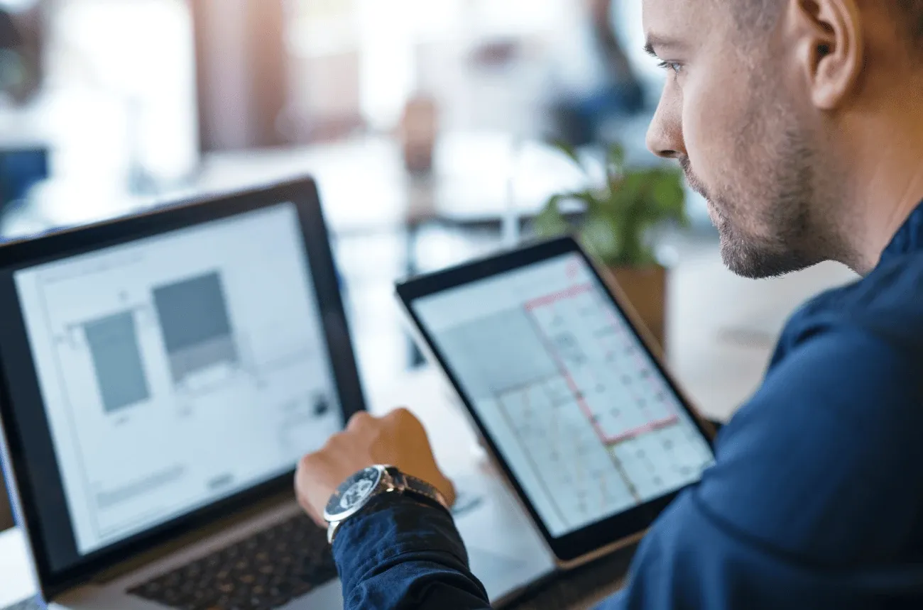 A man in a navy shirt working at a laptop next to a tablet showing a floor plan in an office