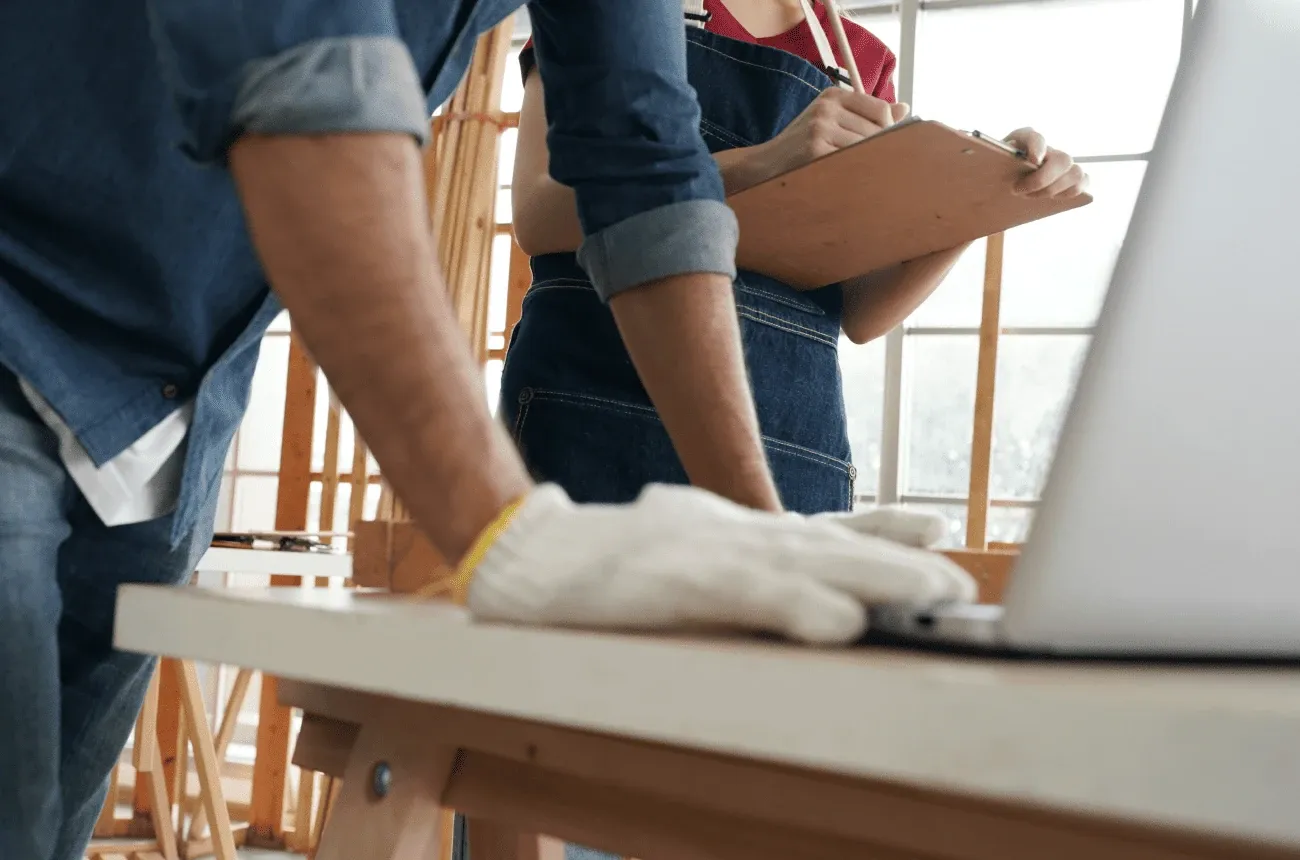 A worker in a denim shirt and white work gloves leaning on a laptop at a workbench while a colleague writes on a clipboard
