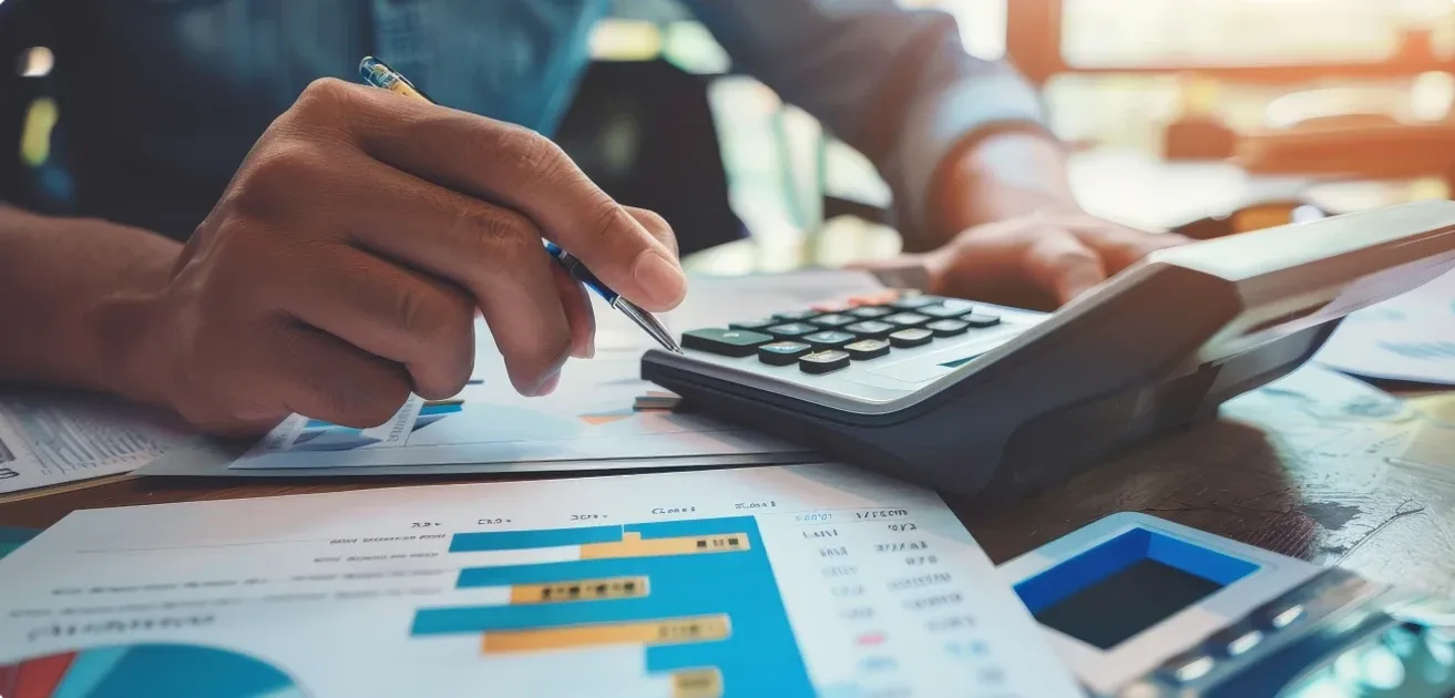 A person holding a pen over printed bar charts while using a calculator at a desk